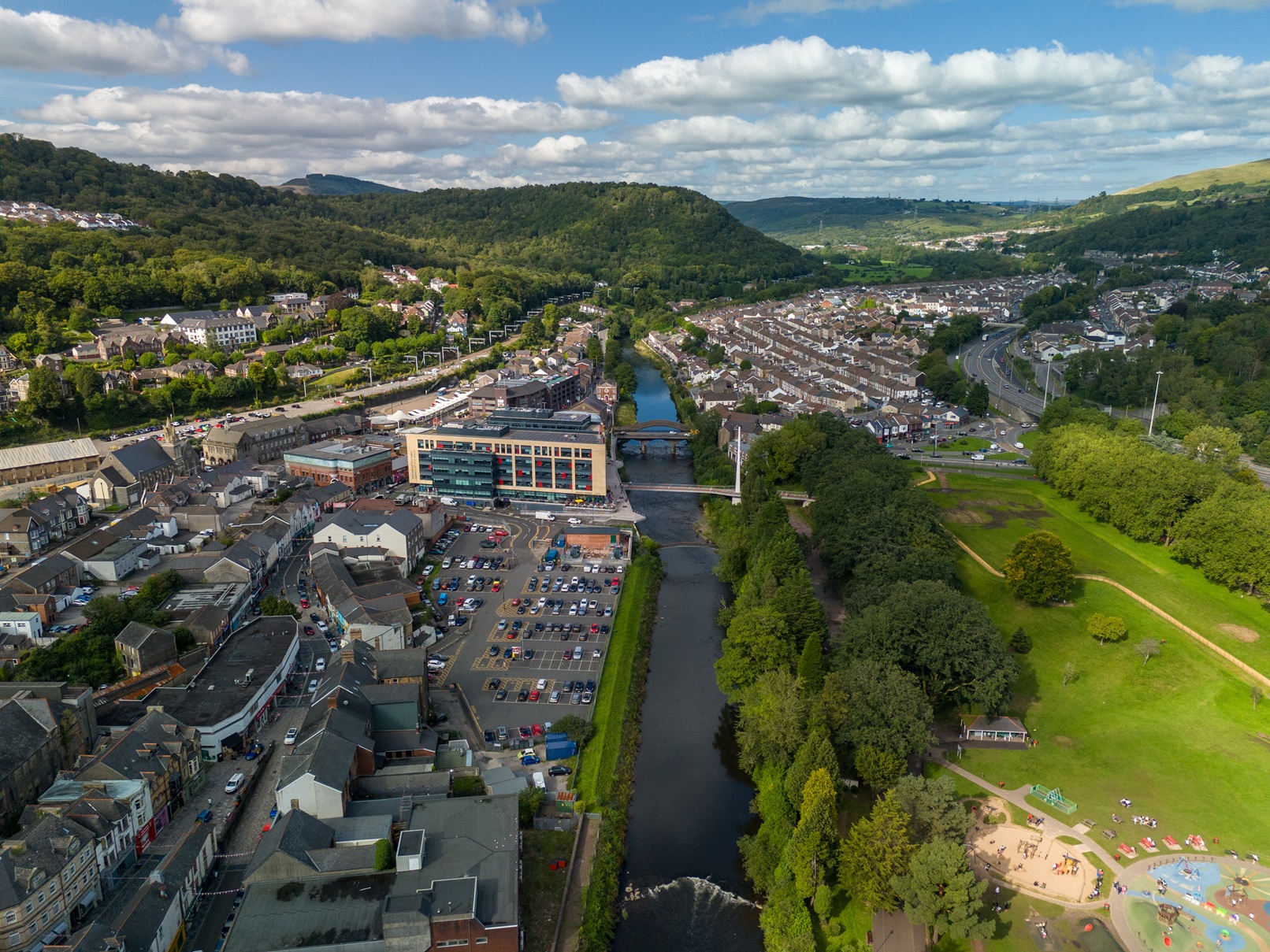 Aerial view of Pontypridd showing the River Taff running through the town centre, with rows of houses on both sides, a modern glass-fronted building and car park near the river, bridges crossing the water, a park with trees and a play area to the right, and green hills in the background under a partly cloudy sky.
